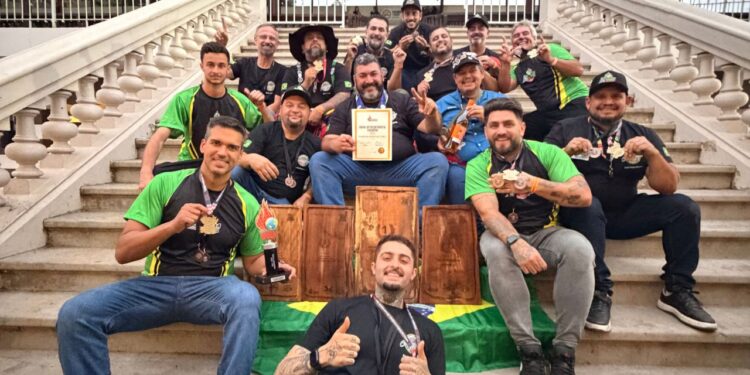 Equipe brasileira campeã mundial de churrasco reunida na escadaria exibindo medalhas, troféus e certificado da conquista.
