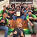 Equipe brasileira campeã mundial de churrasco reunida na escadaria exibindo medalhas, troféus e certificado da conquista.